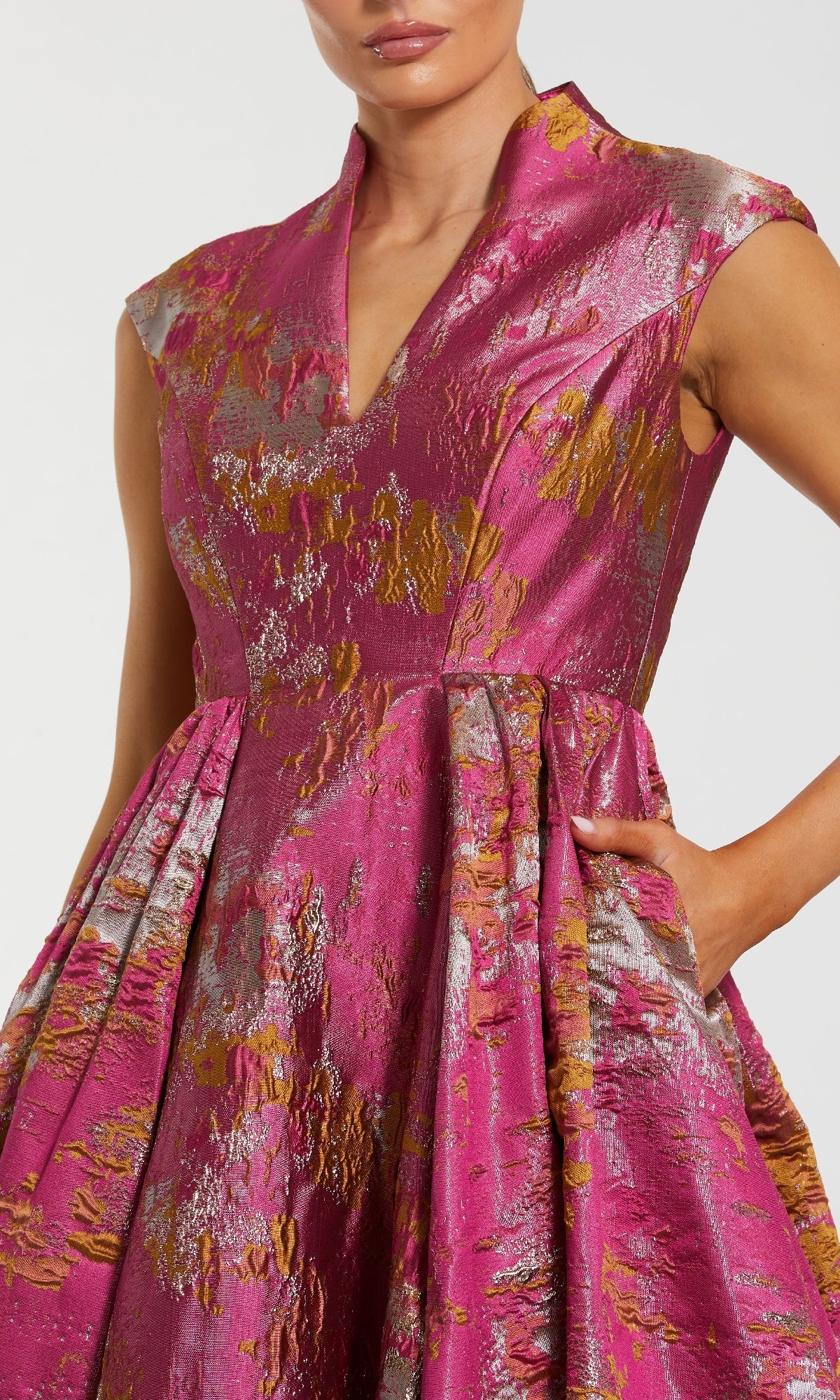 A woman wears the Mac Duggal 68956 long formal dress—a sleeveless pink jacquard gown with gold and silver metallic accents, V-neckline, A-line silhouette, full skirt, and a pocket where she places one hand.
